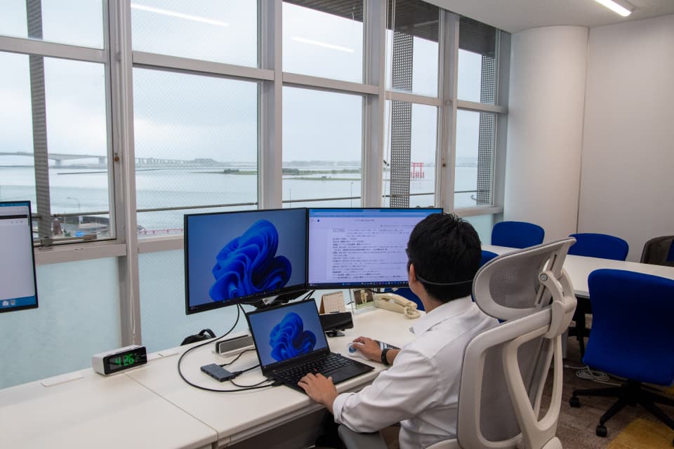 浜名湖を望むオフィスで、デュアルモニターとノートパソコンに向かって開発作業をしている様子